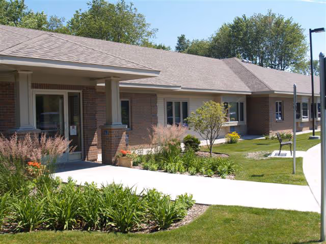 Single-story brick building with a covered entrance, landscaped beds, and a curved concrete walkway in front.