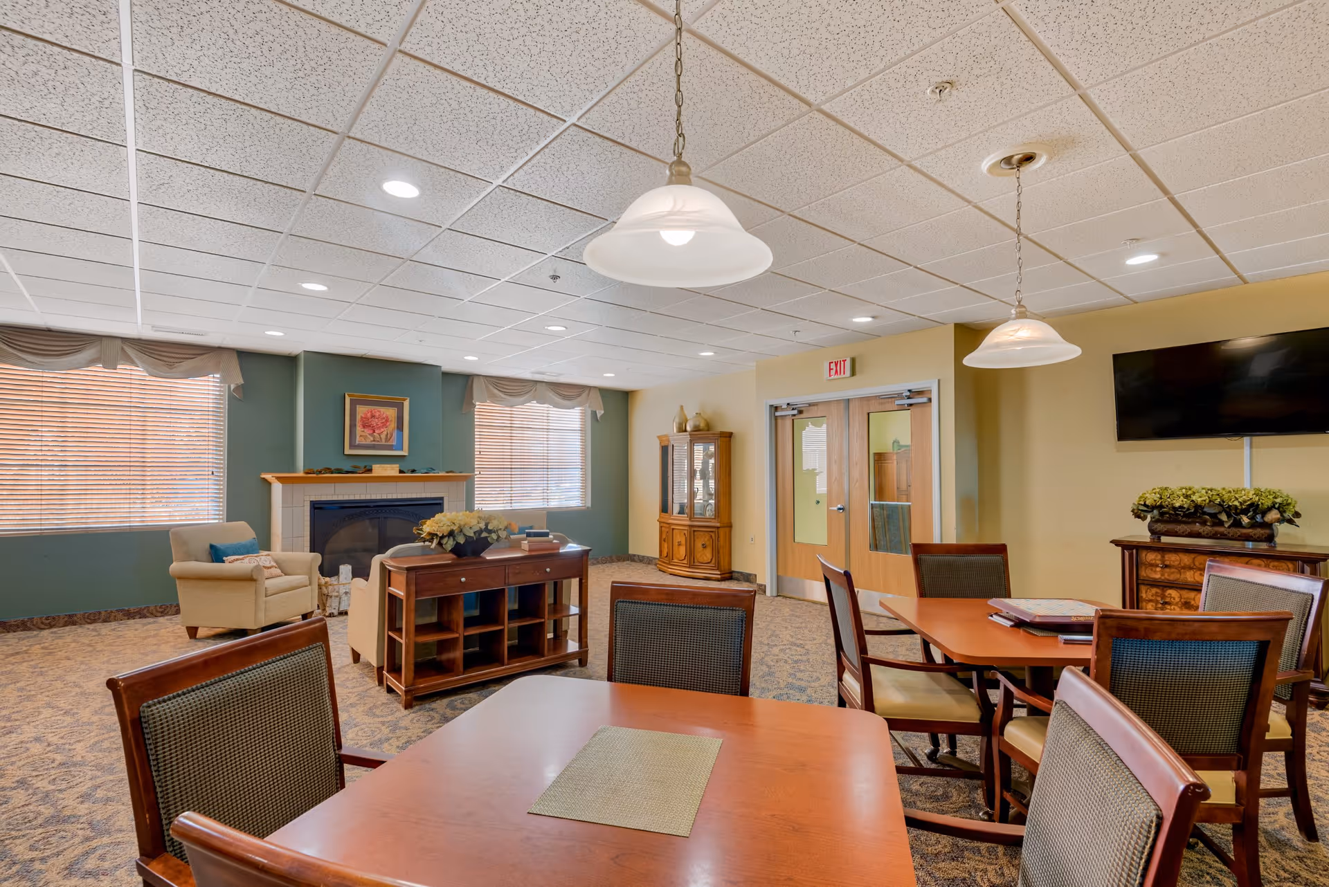 A cozy common area with wooden tables and chairs, a fireplace with a framed picture above it, two armchairs, a cabinet with glass doors, a wall-mounted TV, and two hanging pendant lights. The room has carpeted floors, large windows with blinds, and double doors with an exit sign above them.