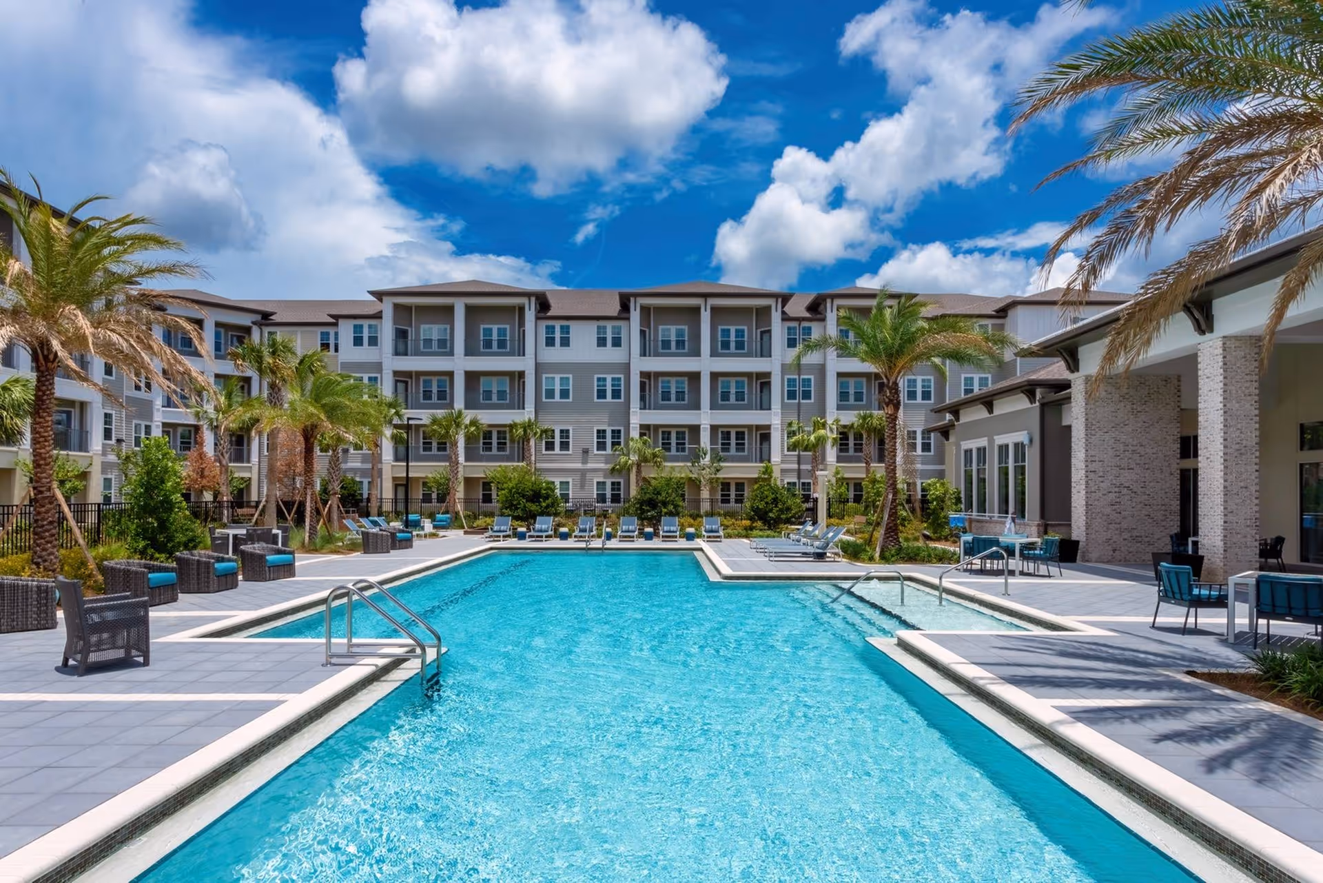 Outdoor swimming pool and seating area in front of a multi-story residential building with palm trees under a blue sky.