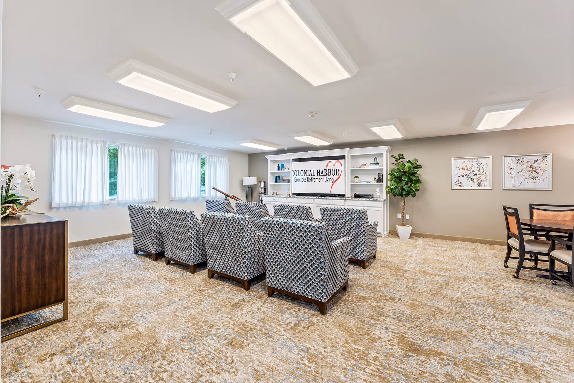 Bright communal lounge with patterned armchairs arranged in rows facing a TV and shelving branded 'Colonial Harbor Gracious Retirement Living'.