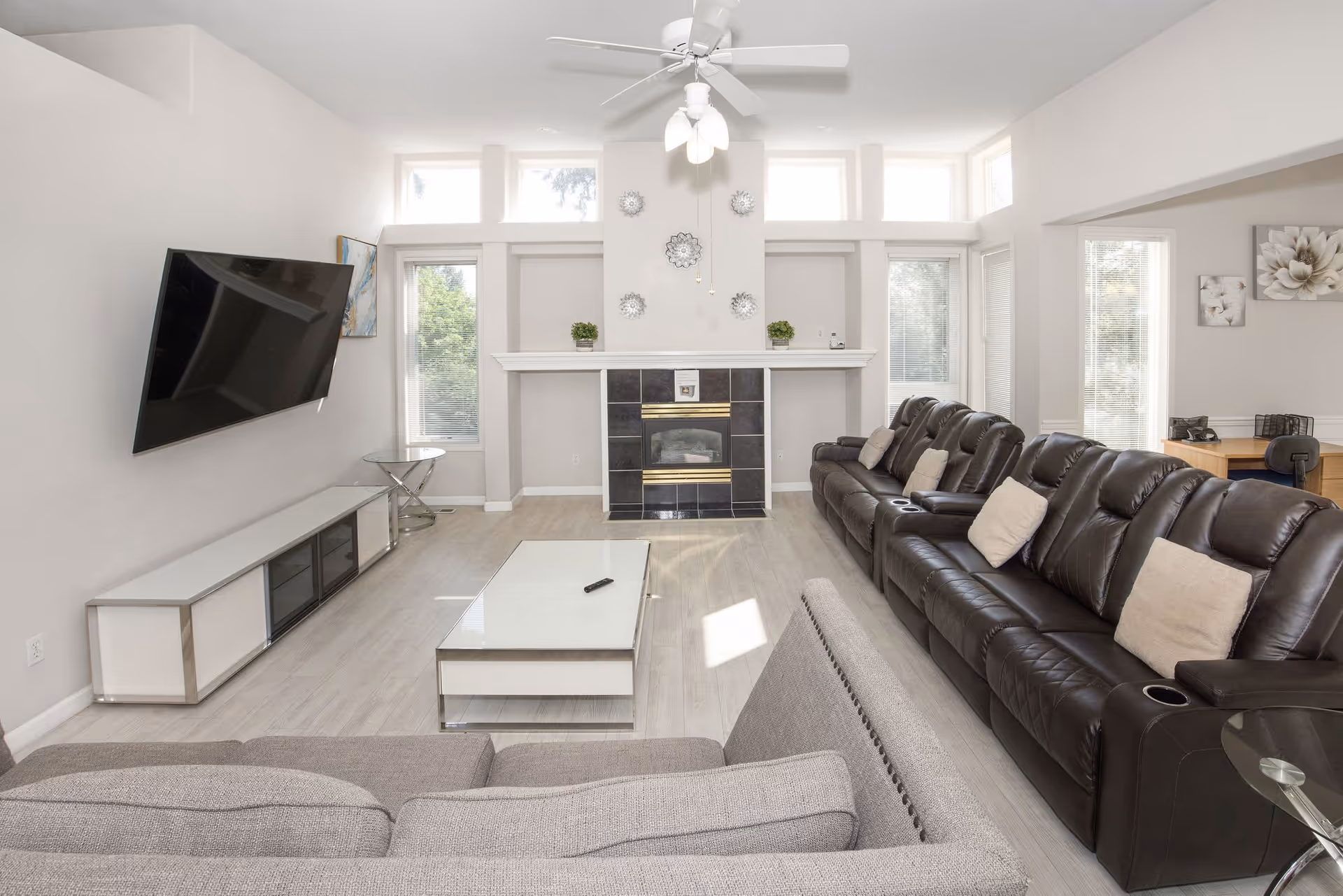 A bright living room with a large dark brown leather sofa with beige pillows, a gray fabric sofa, a white coffee table with a remote control on it, a wall-mounted flat-screen TV, a fireplace with black tiles and decorative wall hangings above it, and several tall windows letting in natural light.