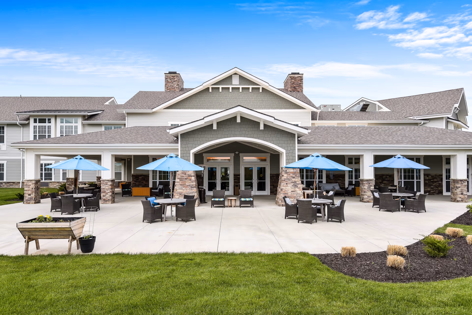 Outdoor patio area of American House Burlington Creek with multiple tables and chairs under blue umbrellas, stone pillars, and a large building entrance in the background under a partly cloudy sky.
