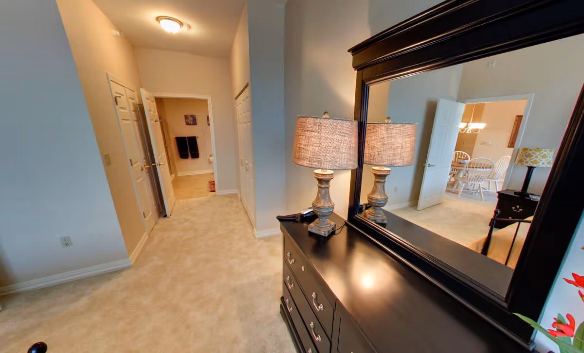 Interior view of a senior living facility room with beige carpet and walls. A black dresser with a large mirror and a lit table lamp is on the right. The mirror reflects a dining area with a table and white chairs. There are multiple doors along the hallway, one leading to a bathroom with towels hanging on the wall.