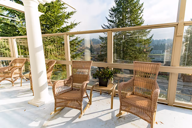 A sunlit outdoor patio area with wicker rocking chairs and a small table holding a potted plant, surrounded by glass railings and overlooking trees and distant hills.