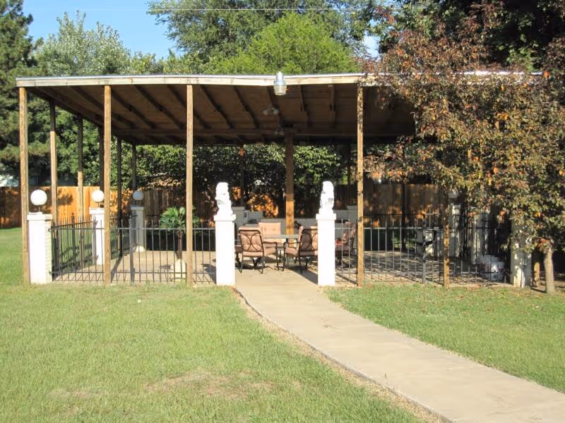 Covered outdoor pavilion with a patio table and chairs under a wooden roof, flanked by white pillars and surrounded by grass and trees.