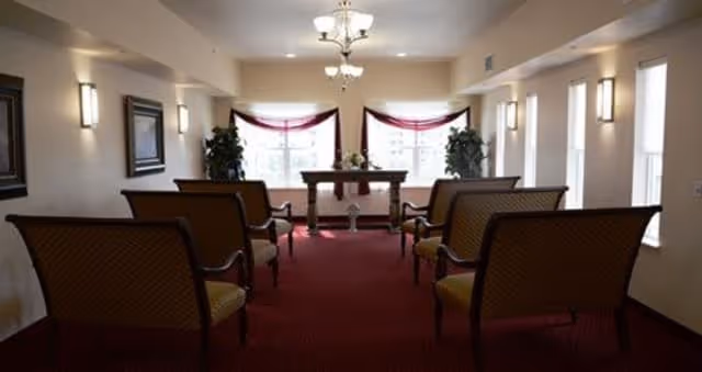 A small chapel or meeting room with rows of cushioned chairs facing a wooden altar table adorned with flowers. The room has red carpet, cream-colored walls, wall-mounted lights, and two large windows with red curtains behind the altar. Two potted plants are placed on either side of the altar.