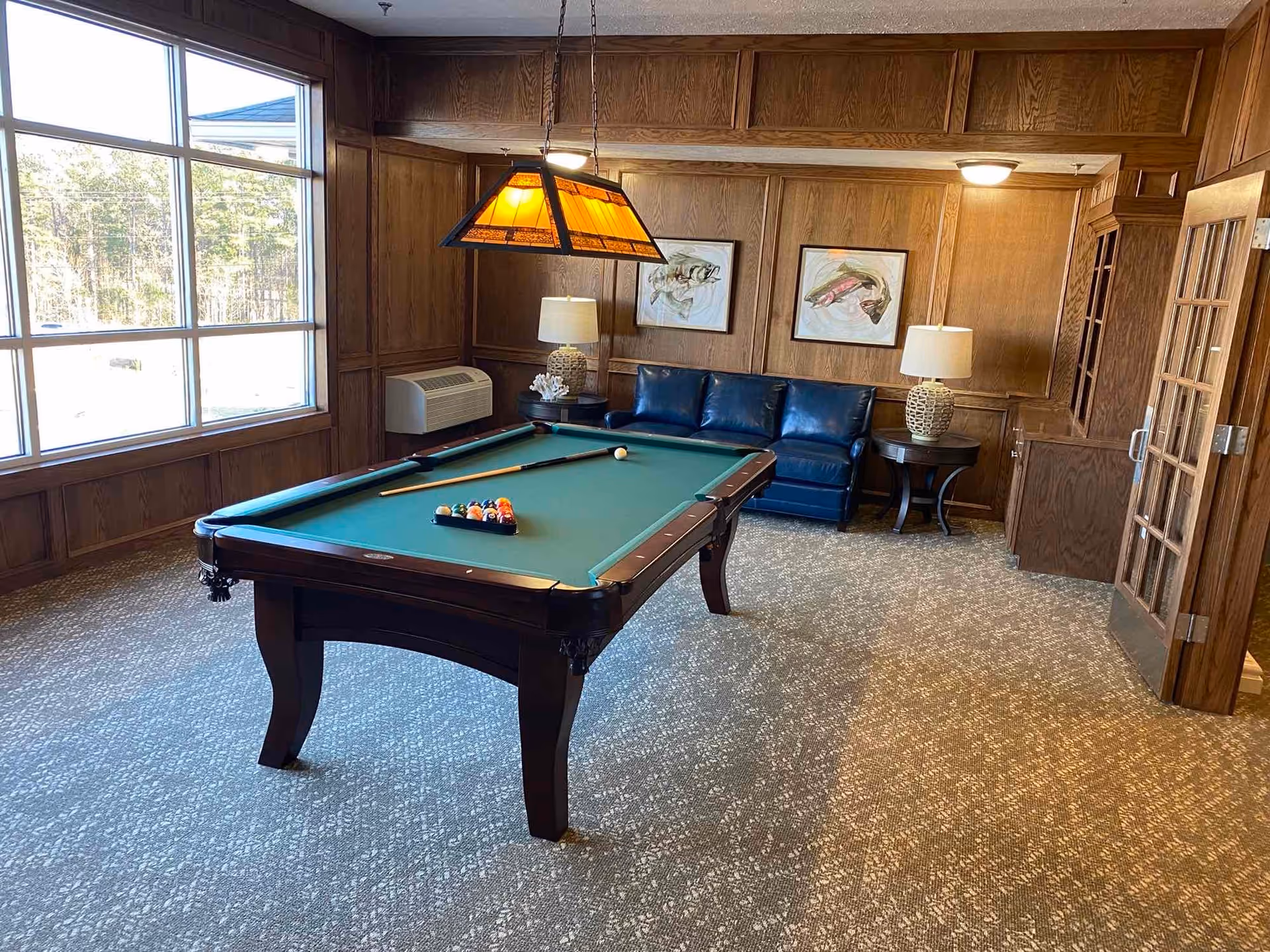 A cozy game room with a pool table in the center, a blue leather couch against a wood-paneled wall, two side tables with lamps, and two framed fish paintings. Large windows let in natural light, and a stained glass pendant light hangs above the pool table.