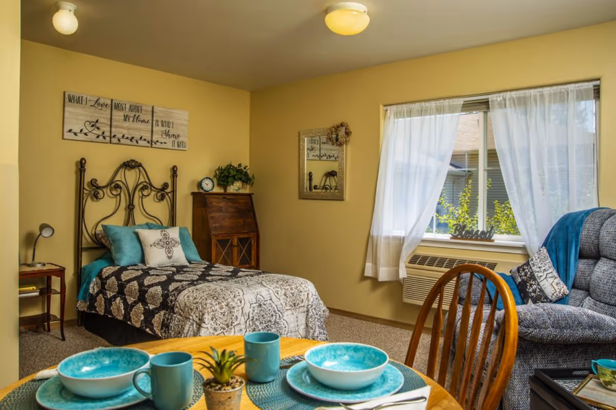 Sunny studio-style senior apartment with a decorated twin bed, dining table set, armchair, and a window with sheer curtains.