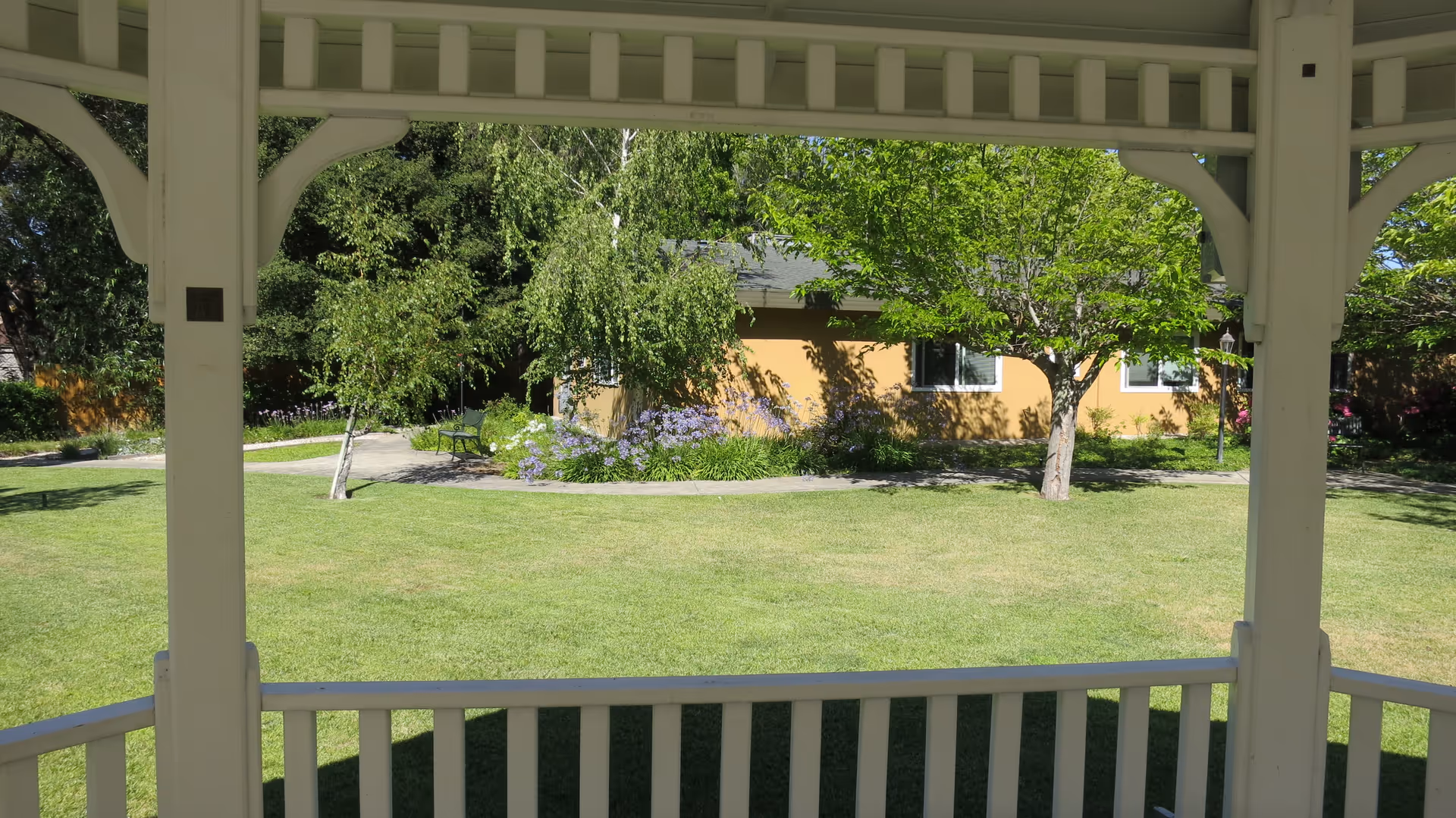 View from inside a white wooden gazebo looking out onto a well-maintained green lawn with trees, bushes, and a yellow building with windows in the background under a clear sky.
