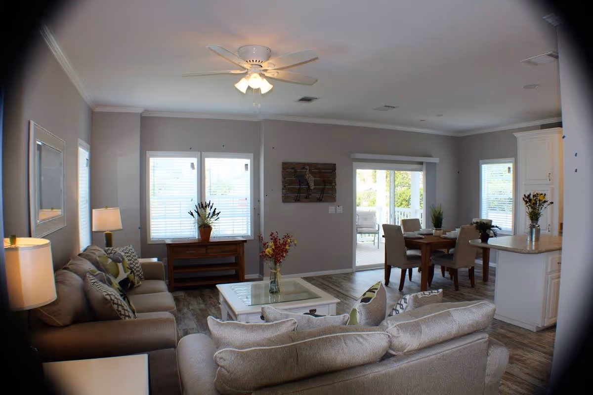 Open-plan living room and dining area in a neutral-toned manufactured home with sofas, coffee table, dining table, and sliding glass door to a patio.