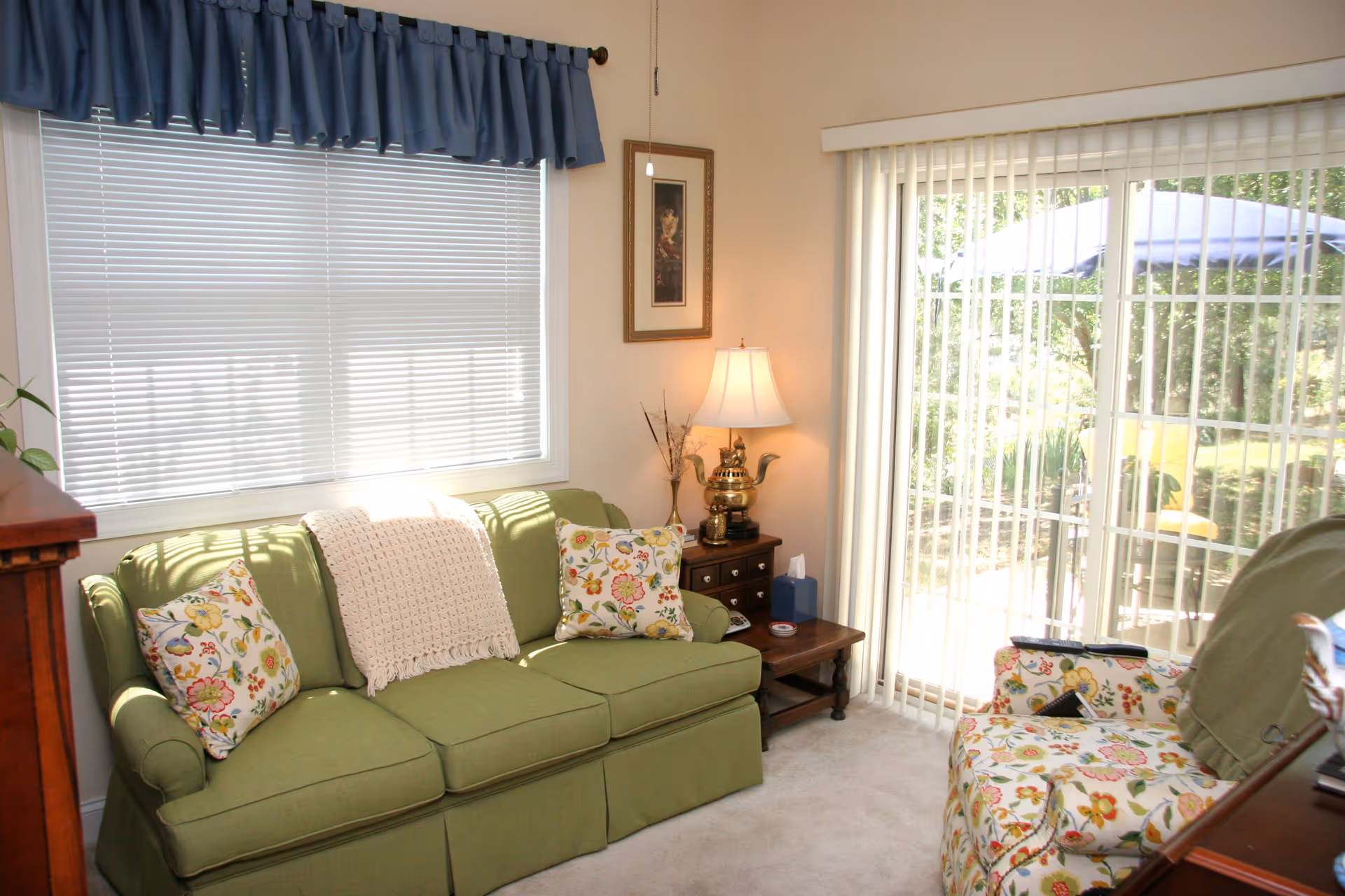 A cozy living room with a green sofa adorned with floral pillows and a white knitted throw. Next to the sofa is a wooden side table with a decorative lamp and a tissue box. A floral armchair is positioned nearby. The room has a large window with closed blinds and a blue valance, and a sliding glass door with vertical blinds that opens to an outdoor patio with a table and umbrella.