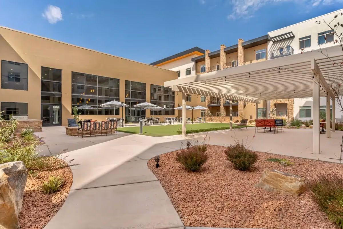 Outdoor courtyard area of a senior living facility with paved walkways, landscaped rock beds with shrubs, seating areas with chairs and umbrellas, and a large white pergola providing shade. The building surrounding the courtyard has large windows and balconies under a clear blue sky.