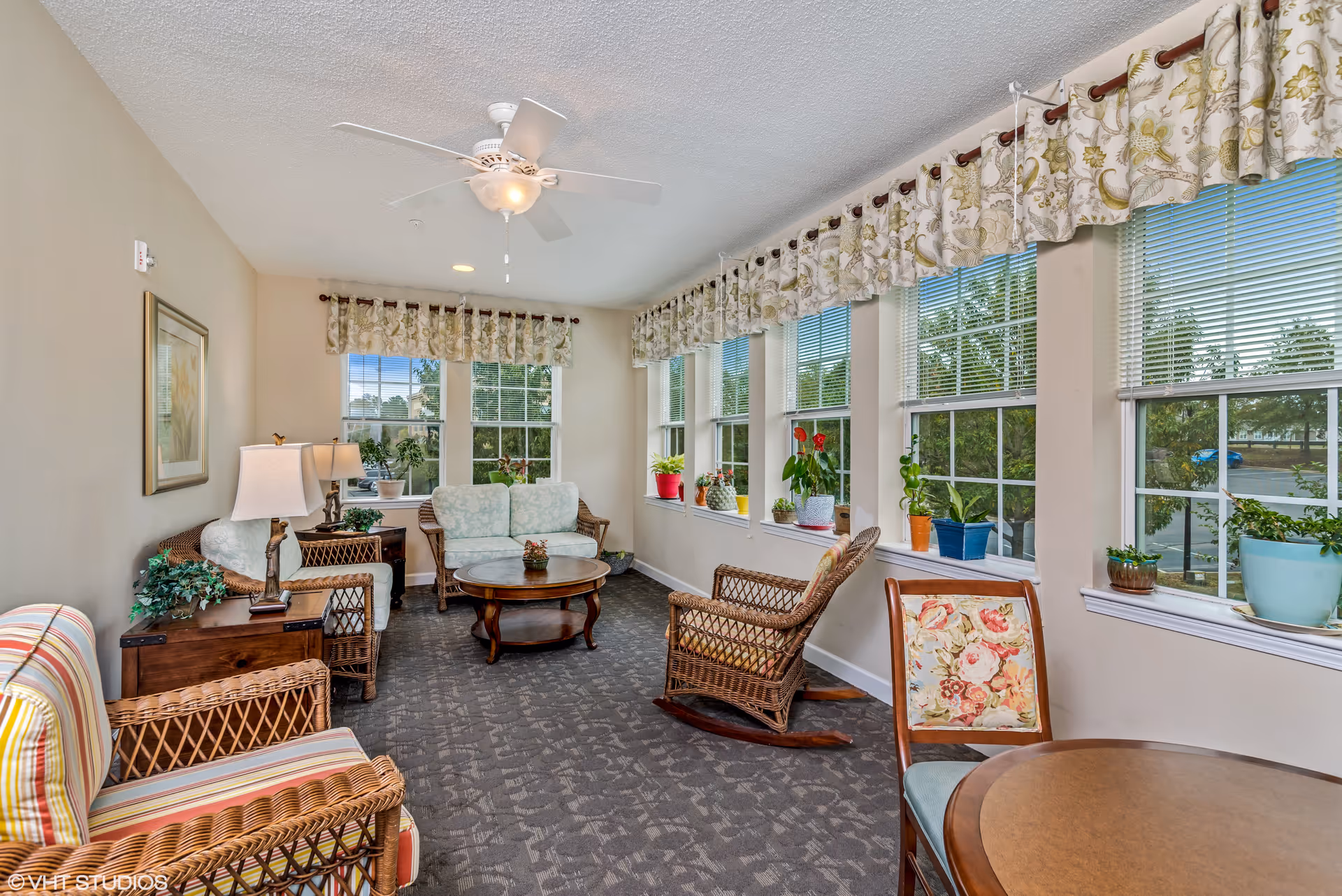 Bright sunroom-style sitting area with wicker chairs, a small sofa, plants on the windowsills, and a ceiling fan.