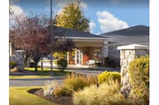 Front entrance of a senior living facility with a covered porch, seating, and landscaped grounds under a partly cloudy sky.