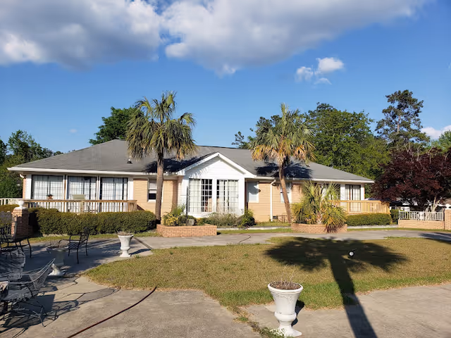 Single-story brick assisted living building with a central entrance, palm trees, and a paved courtyard with planters.