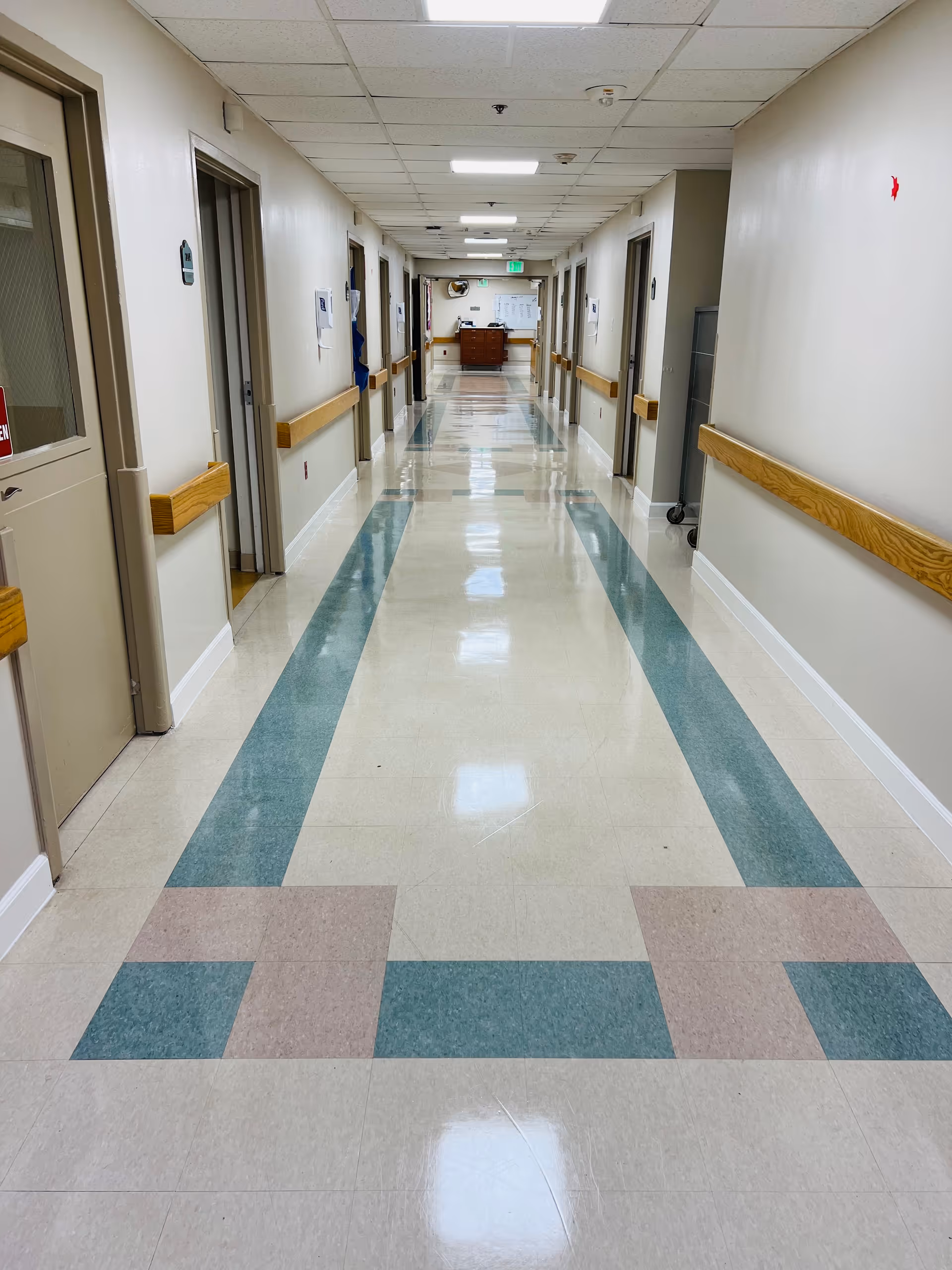 A long, clean hallway in a rehabilitation center with beige walls, handrails on both sides, multiple closed doors, and a shiny tiled floor with a green and beige pattern. The ceiling has recessed lighting panels.