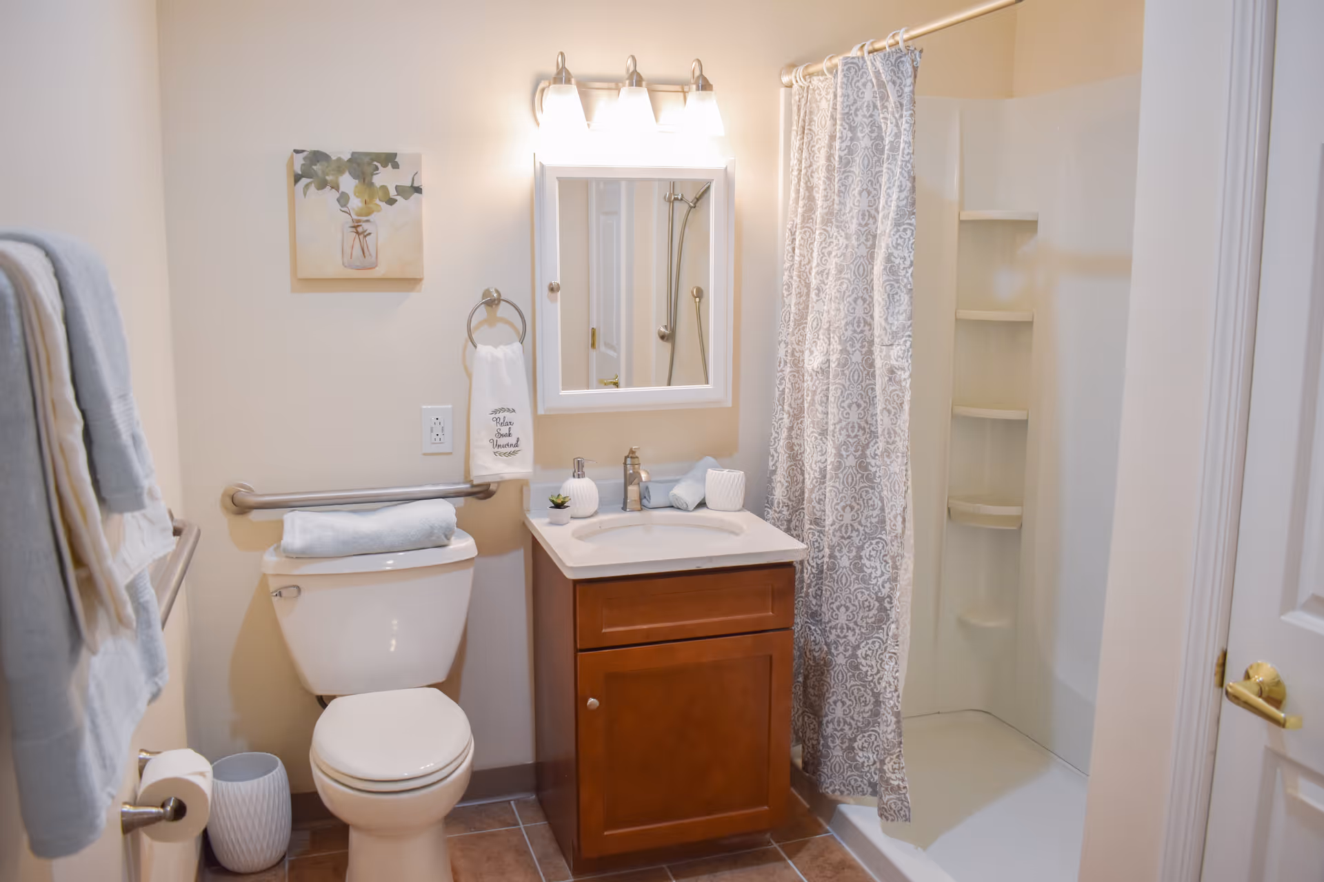 A clean and well-lit bathroom featuring a white toilet with a folded towel on top, a wooden vanity with a sink and mirror above it, a shower with a patterned curtain, and various towels hanging on racks. The walls are light-colored and there is a small framed artwork above the toilet.
