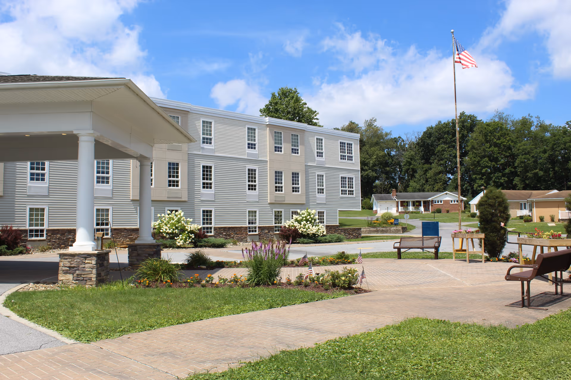 Front entrance and landscaped courtyard of a senior living building with benches and an American flag.