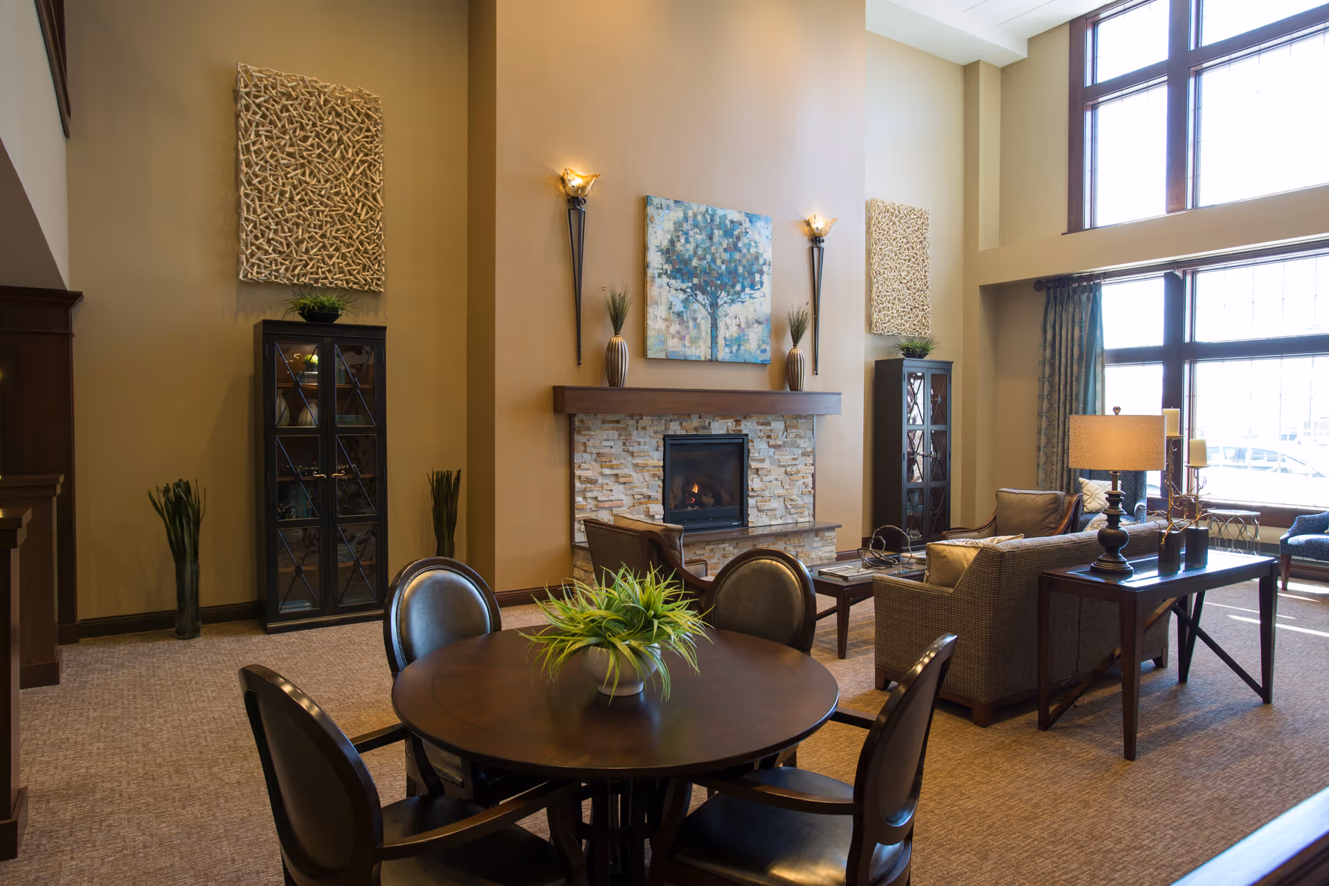 A cozy living room area with a round wooden table and four chairs in the foreground, a stone fireplace with a painting of a tree above it, two tall decorative cabinets on either side, and large windows letting in natural light. The room is decorated with plants, lamps, and wall art.