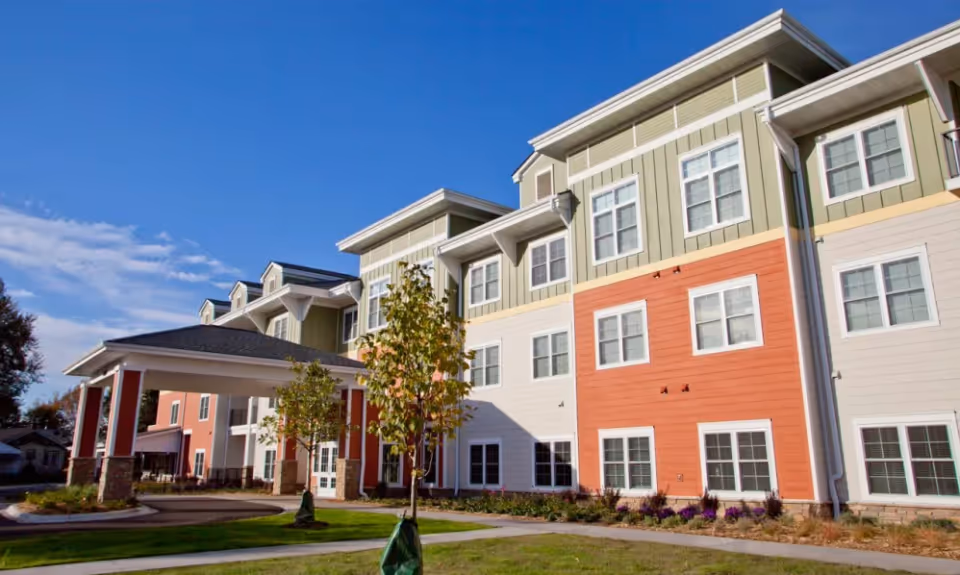 Exterior view of a multi-story senior living facility building with a covered entrance, colorful siding in shades of green, orange, and white, and several windows. There are small trees and landscaped grass areas in front of the building under a clear blue sky.