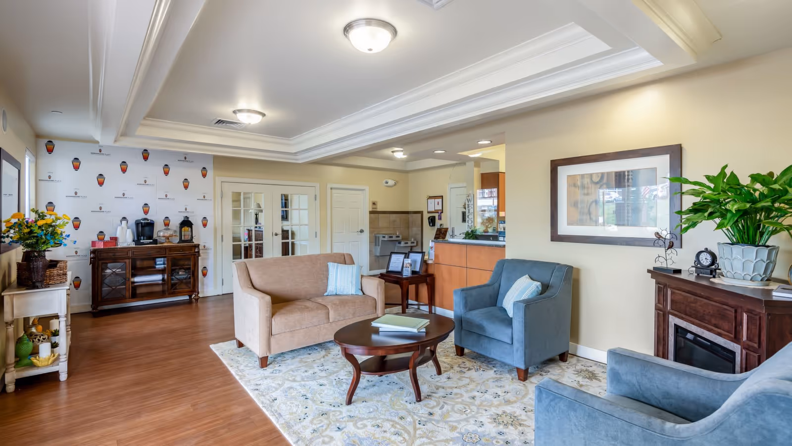 Bright furnished common living area with sofas, armchairs, a coffee table, and a reception desk in a senior living facility.