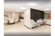Interior view of a senior living facility showing a modern common area with a small kitchenette and dining table with chairs on the left, and a computer workstation with two red stools on the right. The space has light-colored flooring, white walls, and ceiling lights.