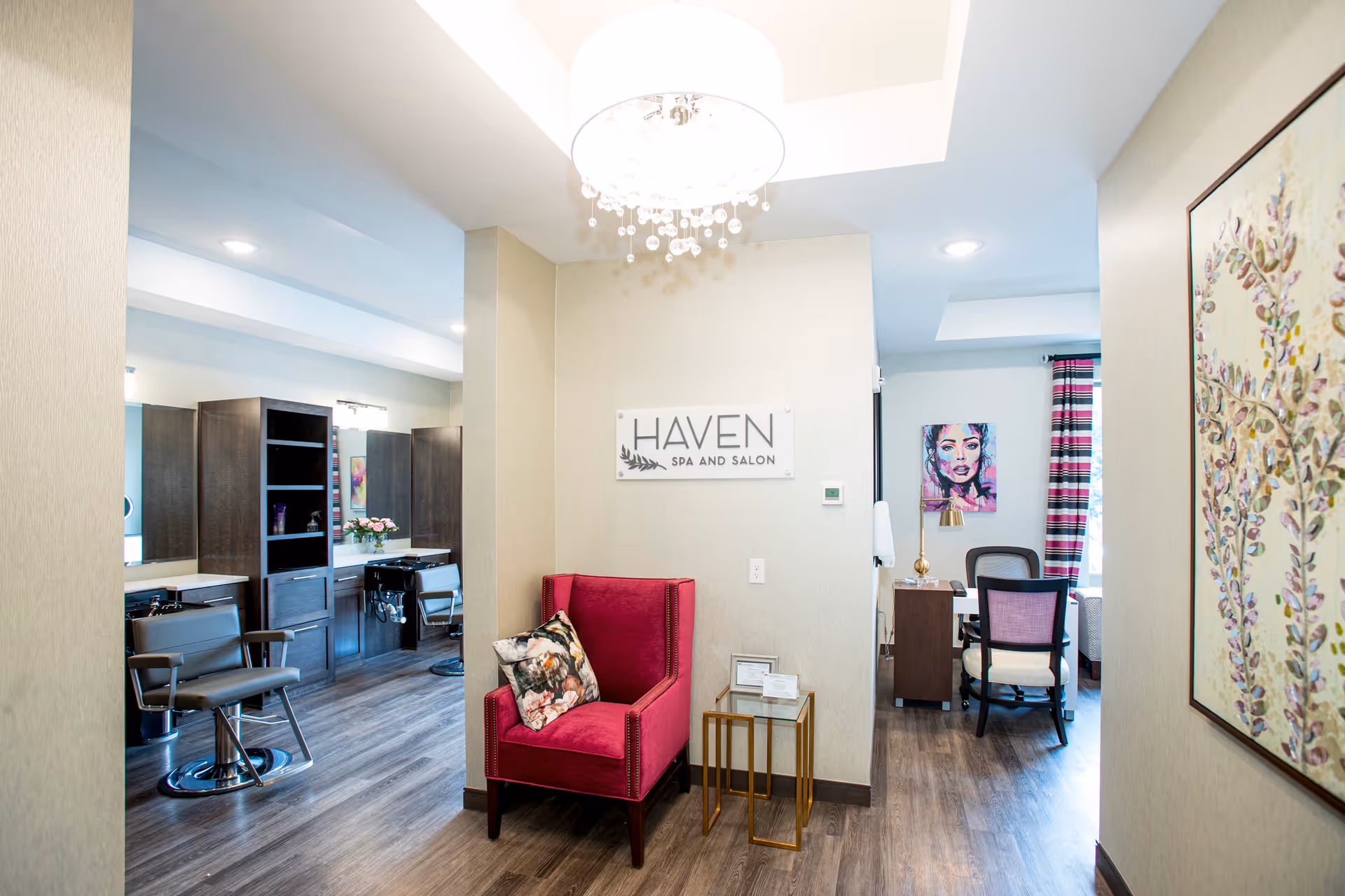 Interior view of a spa and salon area in a senior living facility. The space features a red armchair with a decorative pillow, a small glass side table, and a sign on the wall that reads 'HAVEN SPA AND SALON'. To the left, there are salon chairs and mirrors with dark wood cabinetry. To the right, there is a desk with a chair, a colorful portrait on the wall, and a window with striped curtains. The flooring is wood, and a modern chandelier hangs from the ceiling.