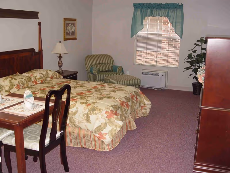 A bedroom in an assisted living facility featuring a bed with floral bedding, a wooden headboard, a small wooden table with two chairs, a striped armchair with matching ottoman, a window with blinds and a green valance, a wooden dresser, a potted plant, and a table lamp on a nightstand.