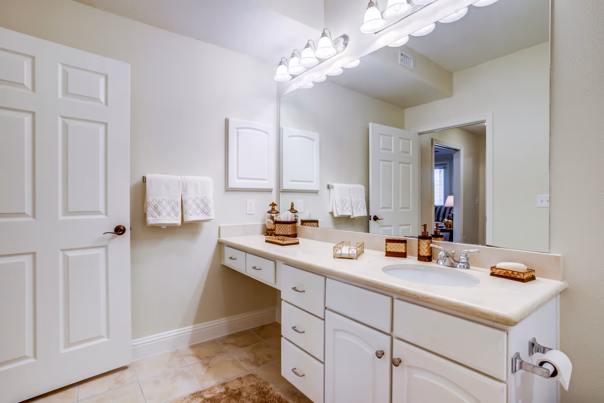A clean and well-lit bathroom vanity area with a large mirror, white cabinets, a beige countertop with a sink, decorative soap dispensers, and neatly folded white towels hanging on the wall. The bathroom door is open, showing a glimpse of an adjacent room with a lamp and window blinds.