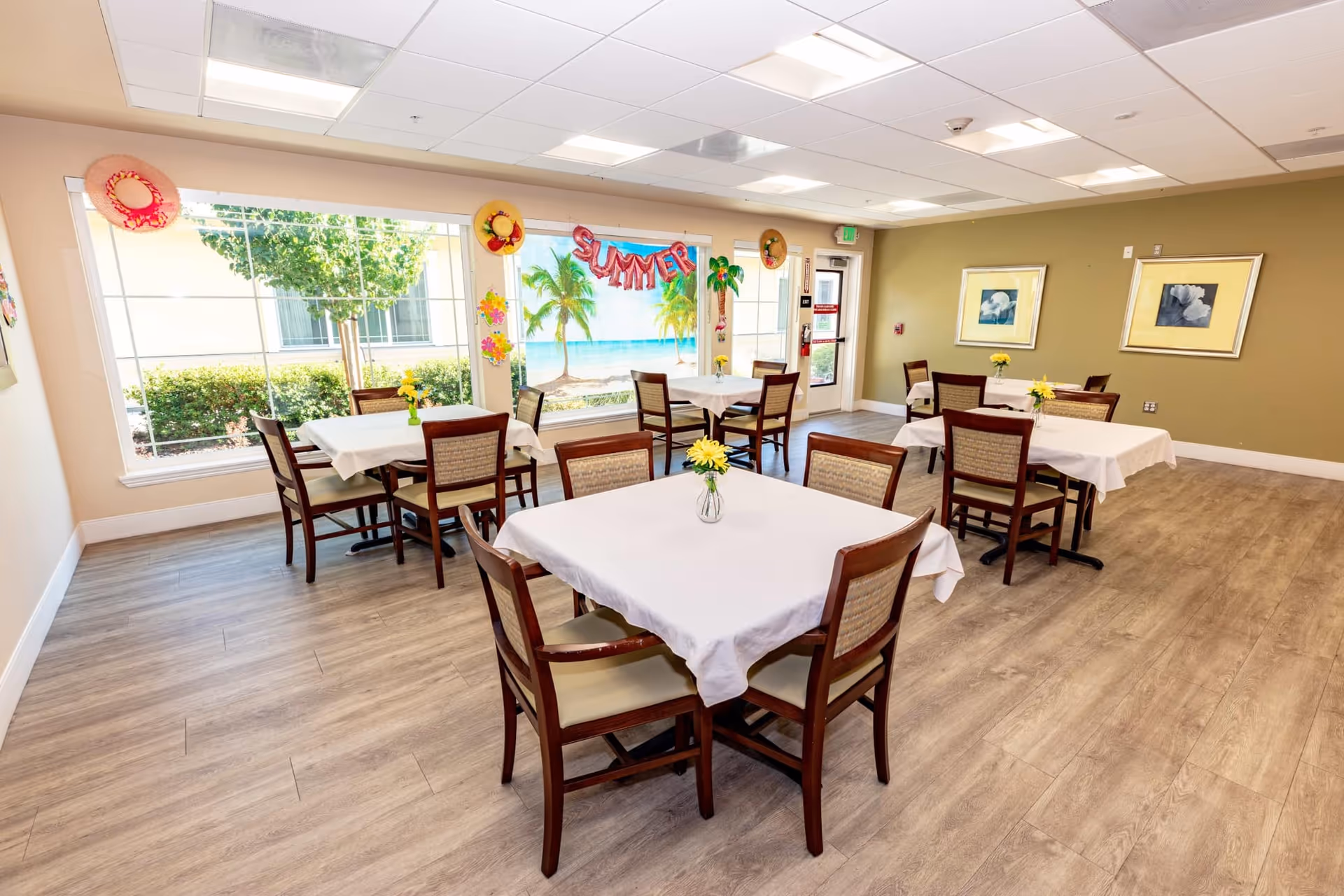 Dining room with multiple tables draped in white cloths, wooden chairs, small yellow flower centerpieces, and a tropical summer mural by a large window.