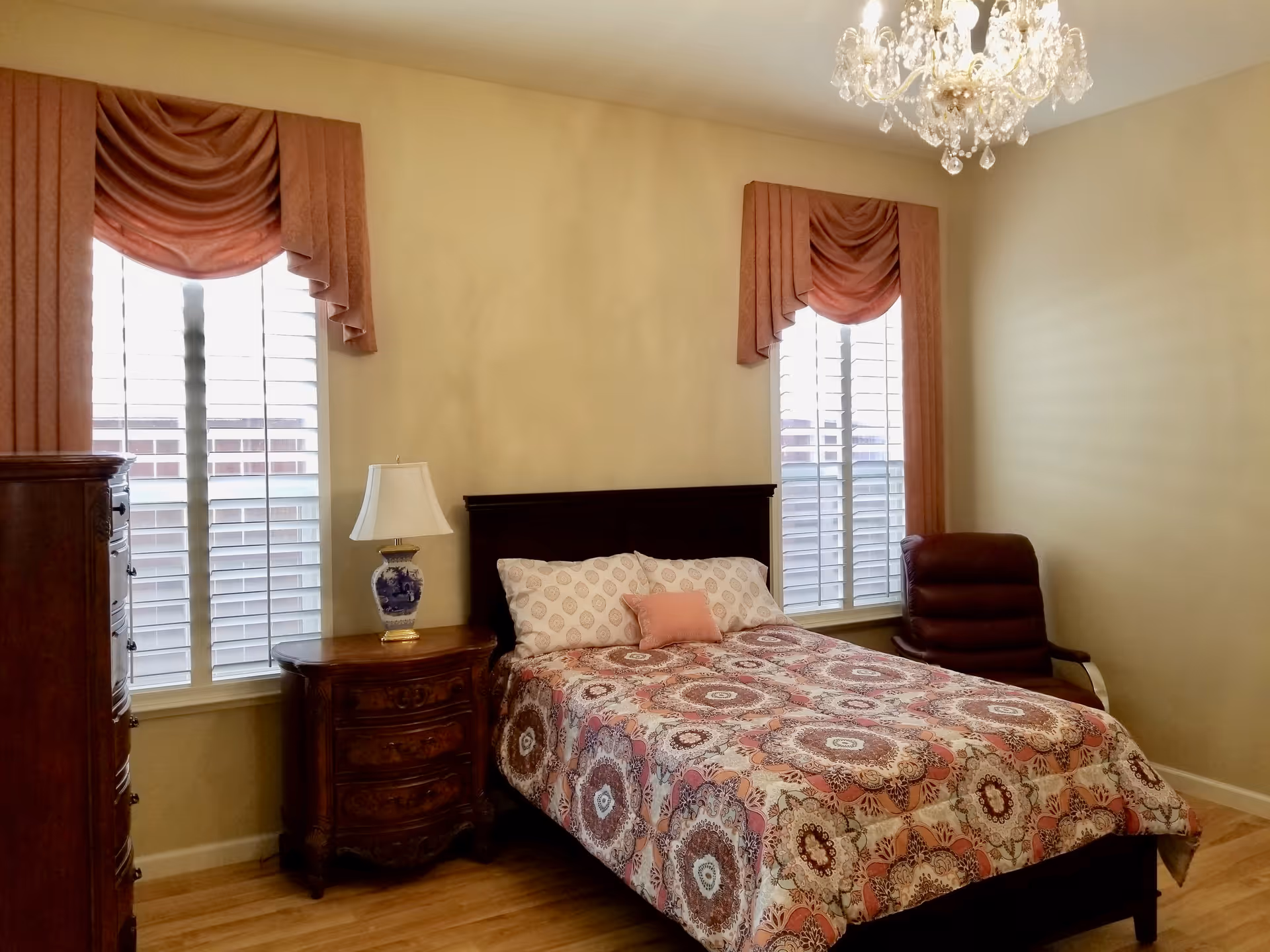 A bedroom with a bed covered in a patterned quilt, two windows with pink drapes and white shutters, a wooden nightstand with a decorative lamp, a wooden dresser, a brown leather chair, and a chandelier hanging from the ceiling.