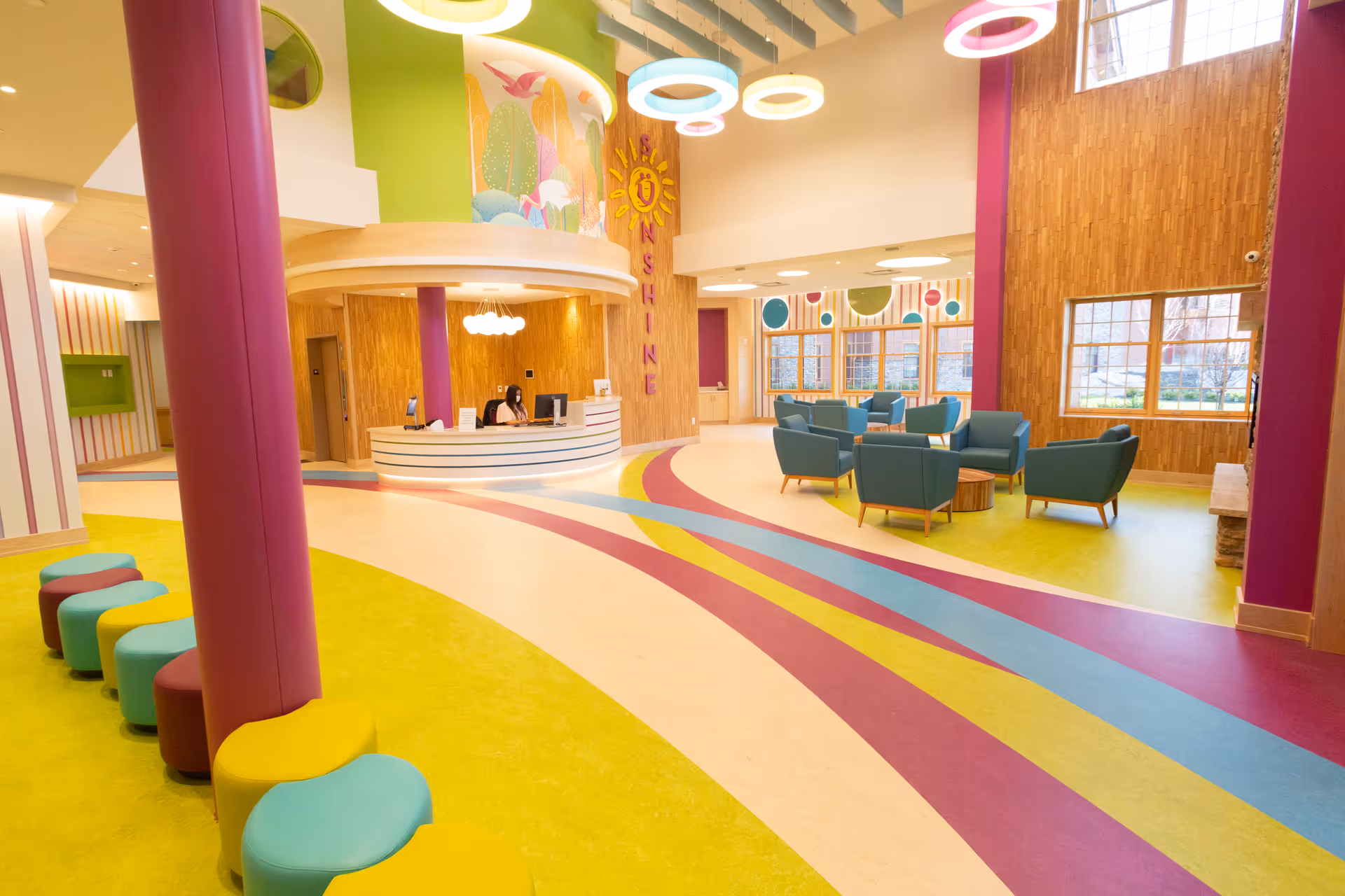 Bright and colorful interior of a children's facility with a curved reception desk, a staff member seated behind it, multicolored circular stools along a wall, modern circular ceiling lights, blue armchairs arranged in a seating area near large windows, and vibrant floor patterns in yellow, blue, and purple.