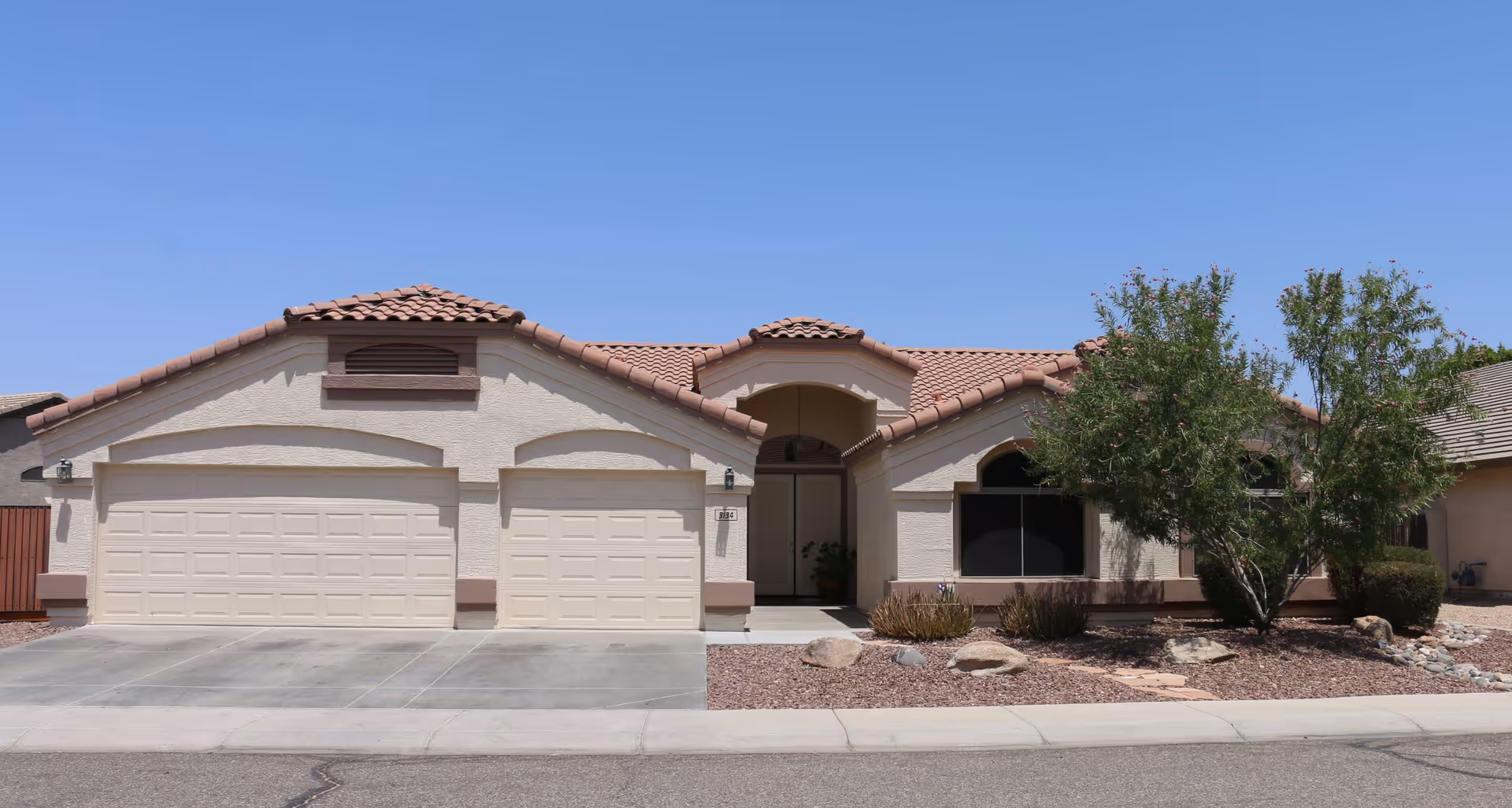 Front exterior view of a single-story residential building with a three-car garage, beige stucco walls, and a red tile roof. There are two trees and some desert landscaping with rocks and shrubs in front of the house under a clear blue sky.