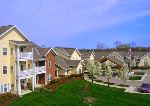 Exterior view of River Terrace Retirement Community showing multiple connected residential buildings with yellow siding and brick accents, white balconies, a covered entrance, landscaped green lawns, blooming trees, and a clear blue sky.