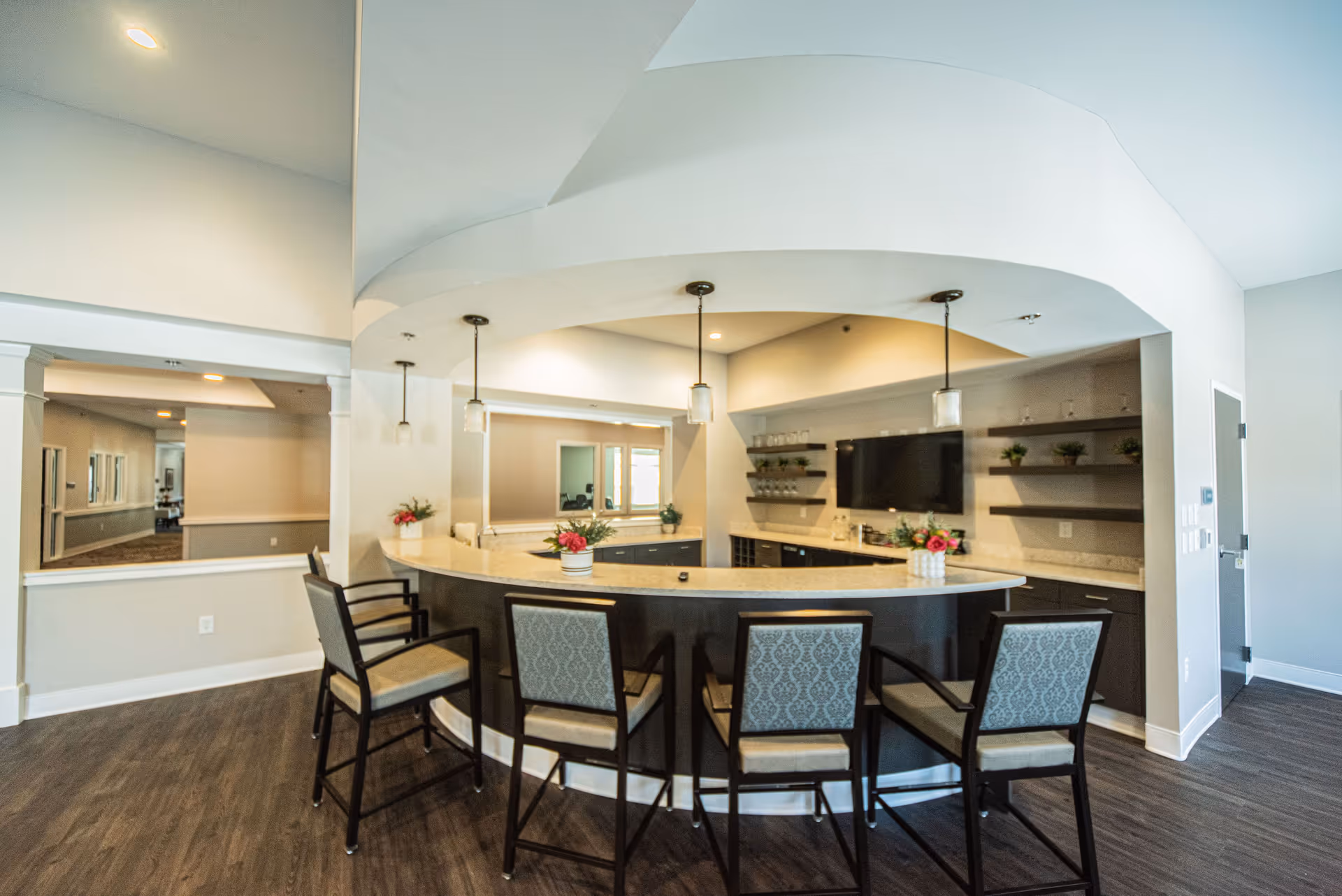 Interior view of a modern common area with a curved bar counter surrounded by four high chairs with patterned upholstery. The bar area has pendant lights hanging from the ceiling, a large mirror on the back wall, shelves with small plants and glassware, and a mounted flat-screen TV. The floor is dark wood, and the walls are painted light gray.