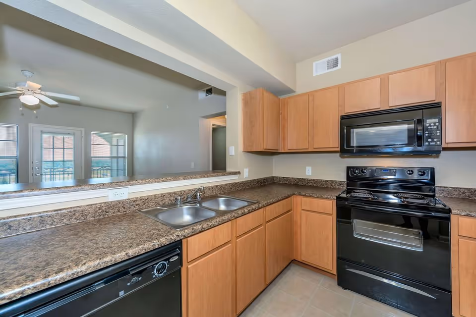 Modern kitchen with light wood cabinets, black electric stove and microwave, double stainless steel sink, and granite countertops. The kitchen opens to a living area with a ceiling fan and large windows with blinds.