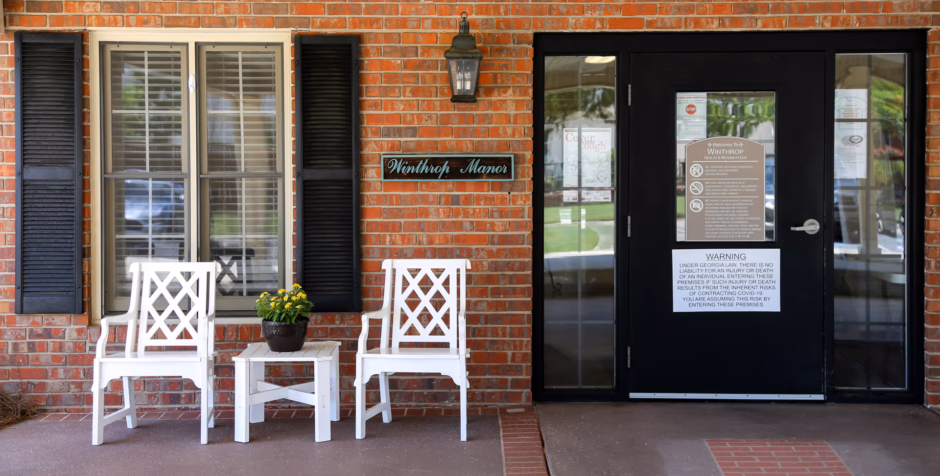 Entrance to Winthrop Manor with a brick exterior wall, two white chairs and a small white table with a potted plant between them, a window with black shutters, and a black door with multiple notices posted on it.