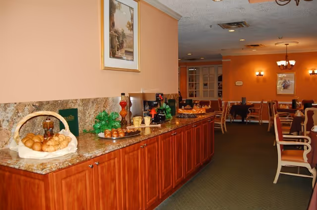 Interior view of a dining area with wooden cabinets topped with granite, displaying baskets of bread rolls, coffee cups, and coffee machines. The room has peach-colored walls, framed artwork, and multiple tables and chairs arranged for dining.