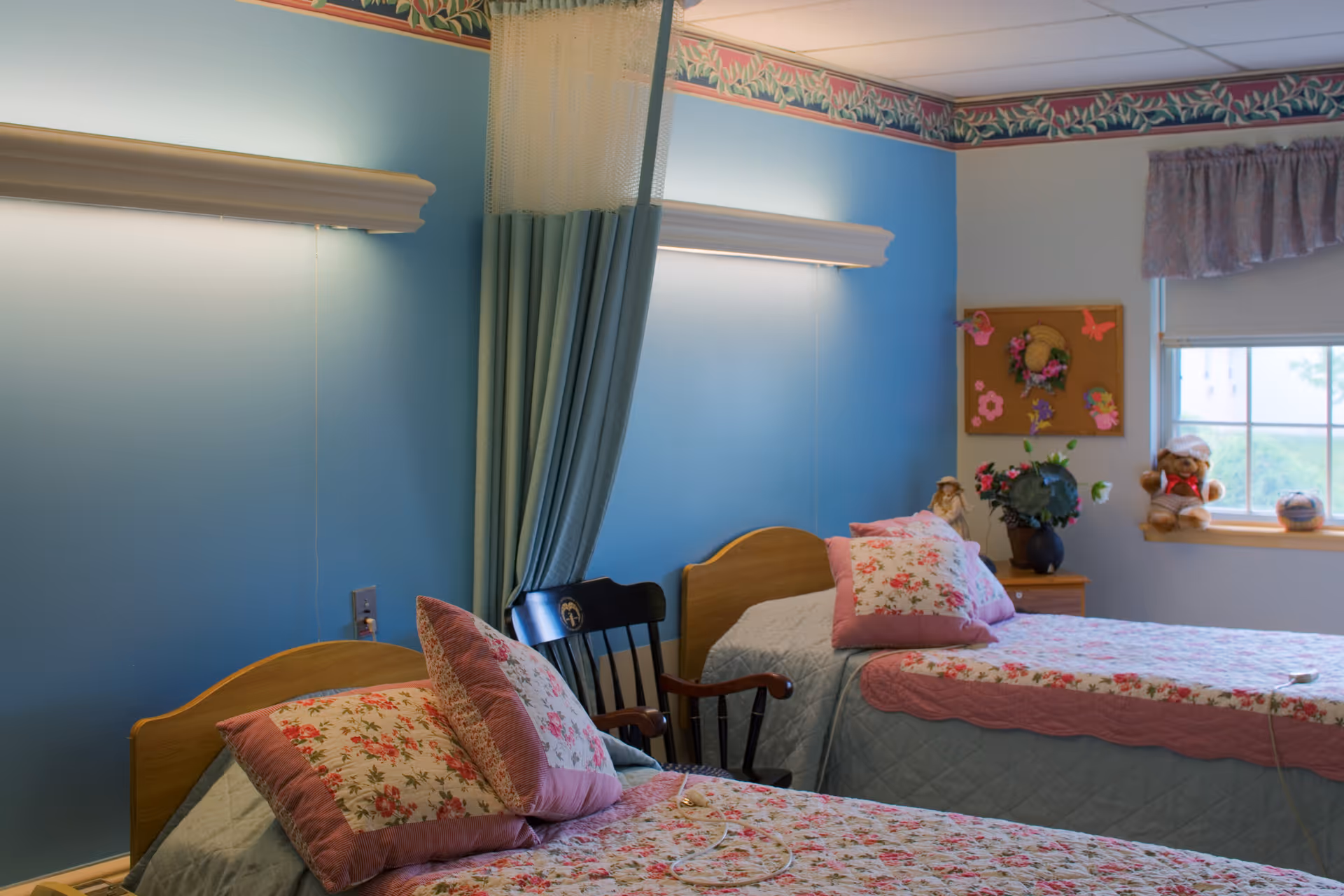 A cozy twin bedroom with two floral quilts on adjacent beds, a window, bedside table with decorations, and a privacy curtain.
