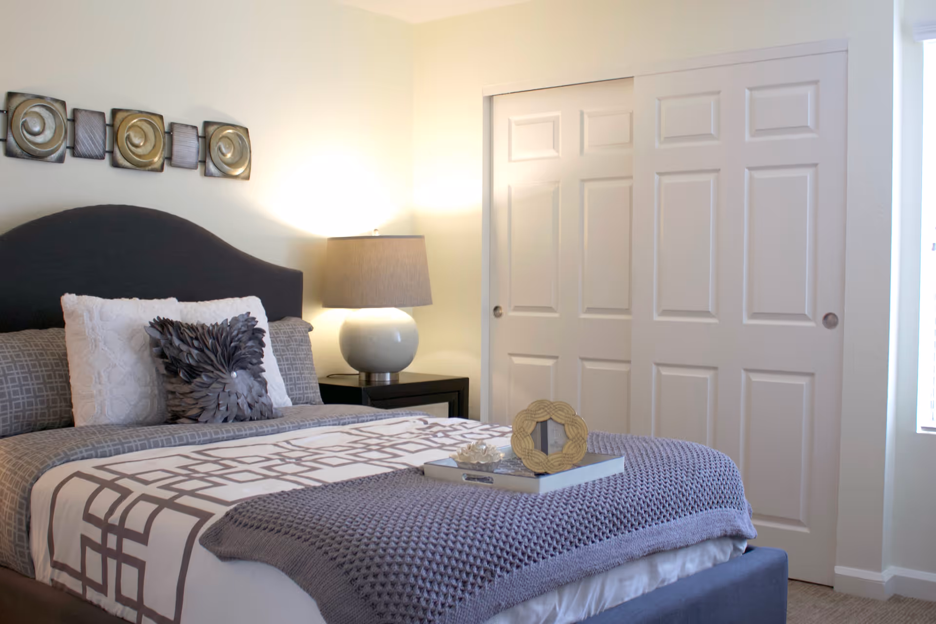 A neatly made bed with a dark upholstered headboard, gray and white patterned bedding, and decorative pillows. A bedside table with a round lamp is next to the bed. Behind the bed, there is a wall decoration with square metallic art pieces. To the right, there are white sliding closet doors and a window letting in natural light.