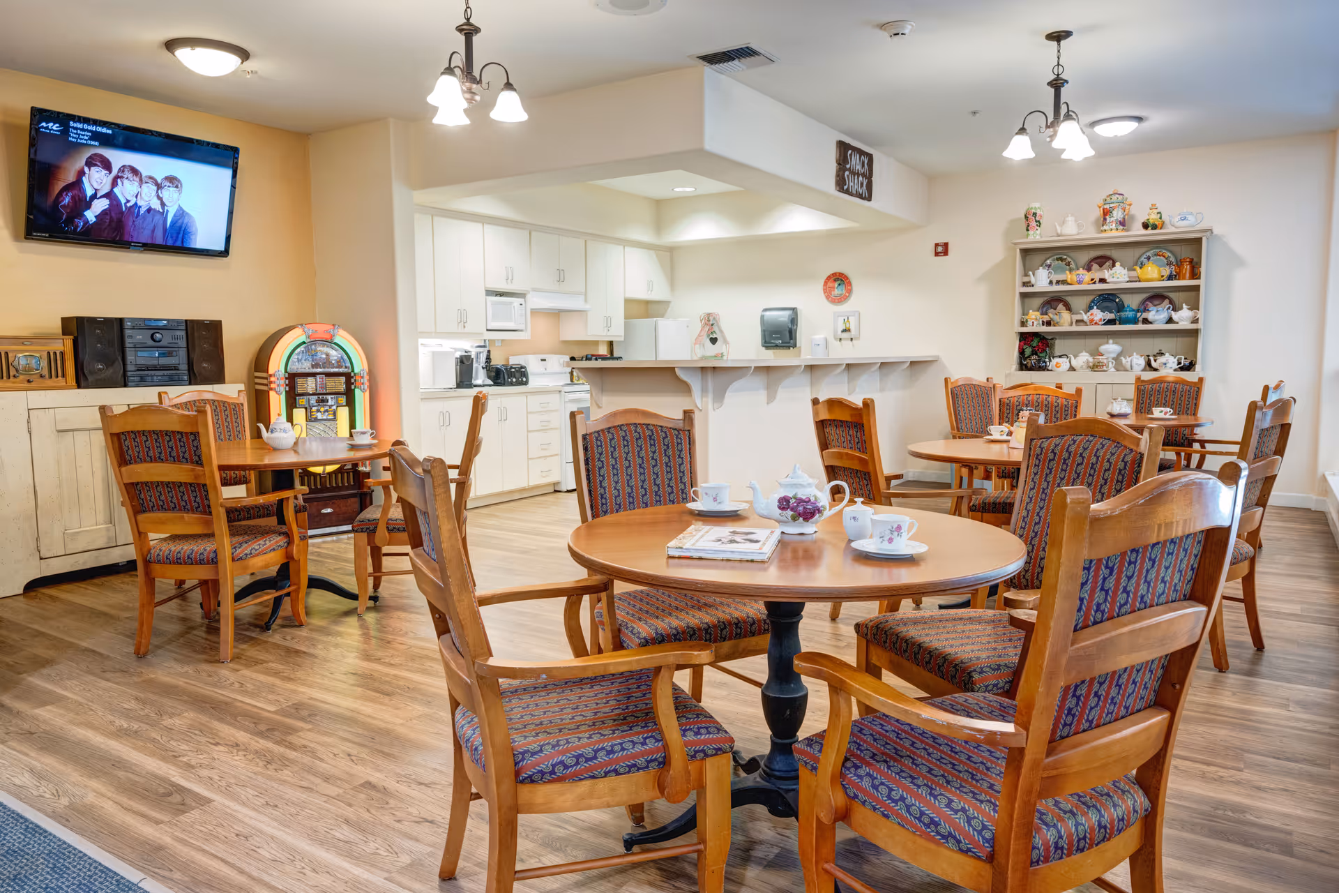 A cozy dining area in a senior living facility with several round wooden tables and cushioned chairs arranged around them. The tables have teapots, cups, and a book on them. In the background, there is a small kitchen area with white cabinets, a microwave, and a refrigerator. A vintage jukebox and a stereo system are placed against a wall with a mounted TV above showing an image of The Beatles. A wooden shelf filled with various teapots and decorative items is also visible. The room has warm lighting and wooden flooring.