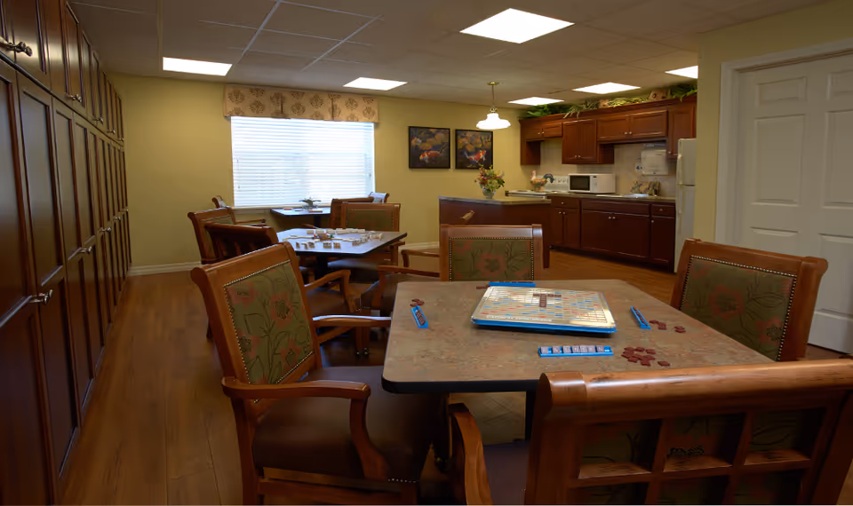 A cozy common area with several tables and chairs, some set up for playing board games like Scrabble. The room features wooden flooring, a large window with blinds and a valance, and a kitchen area with dark wood cabinets, a microwave, and a refrigerator. The walls are painted a warm beige color, and there are two framed paintings on the wall near the kitchen.