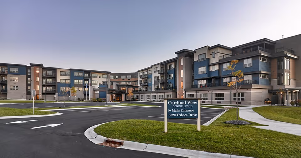 Exterior view of Cardinal View Senior Living facility showing a modern multi-story building with balconies, a paved driveway, green lawns, and a sign indicating the main entrance at 3820 Tribeca Drive.