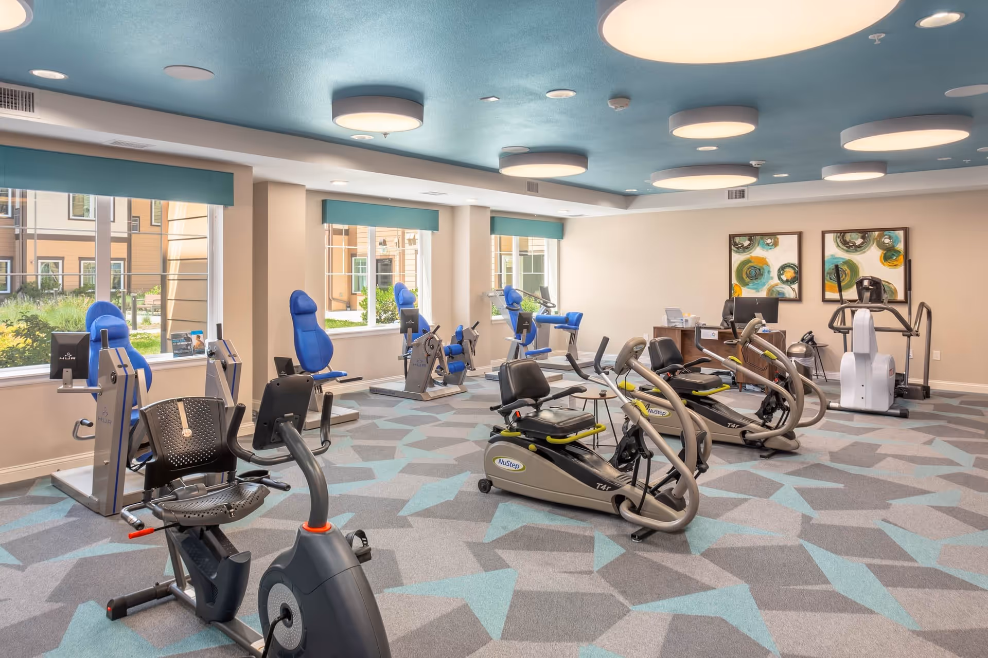 A spacious fitness room with various exercise equipment including recumbent bikes and strength training machines. The room has large windows with teal valances allowing natural light to enter. The ceiling is painted teal with multiple round light fixtures. The floor is carpeted with a geometric pattern in shades of gray and teal. Two abstract paintings hang on the far wall above a small desk with a computer and other office items.