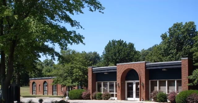 Exterior view of a single-story brick building with large windows and an arched entrance, surrounded by trees and greenery under a clear blue sky.
