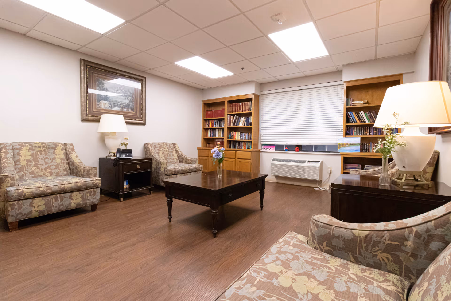 A cozy living room with floral patterned armchairs arranged around a dark wooden coffee table. There are two wooden bookshelves filled with books against the far wall, a window with closed blinds, and two table lamps on dark wooden side tables. The floor is wooden, and a framed painting hangs on the wall.