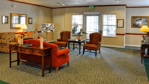 A cozy living room area in Oak Woods Manor featuring patterned wallpaper, a floral sofa, two striped armchairs, a wooden desk with a lamp, and a small table with a plant near large windows letting in natural light.