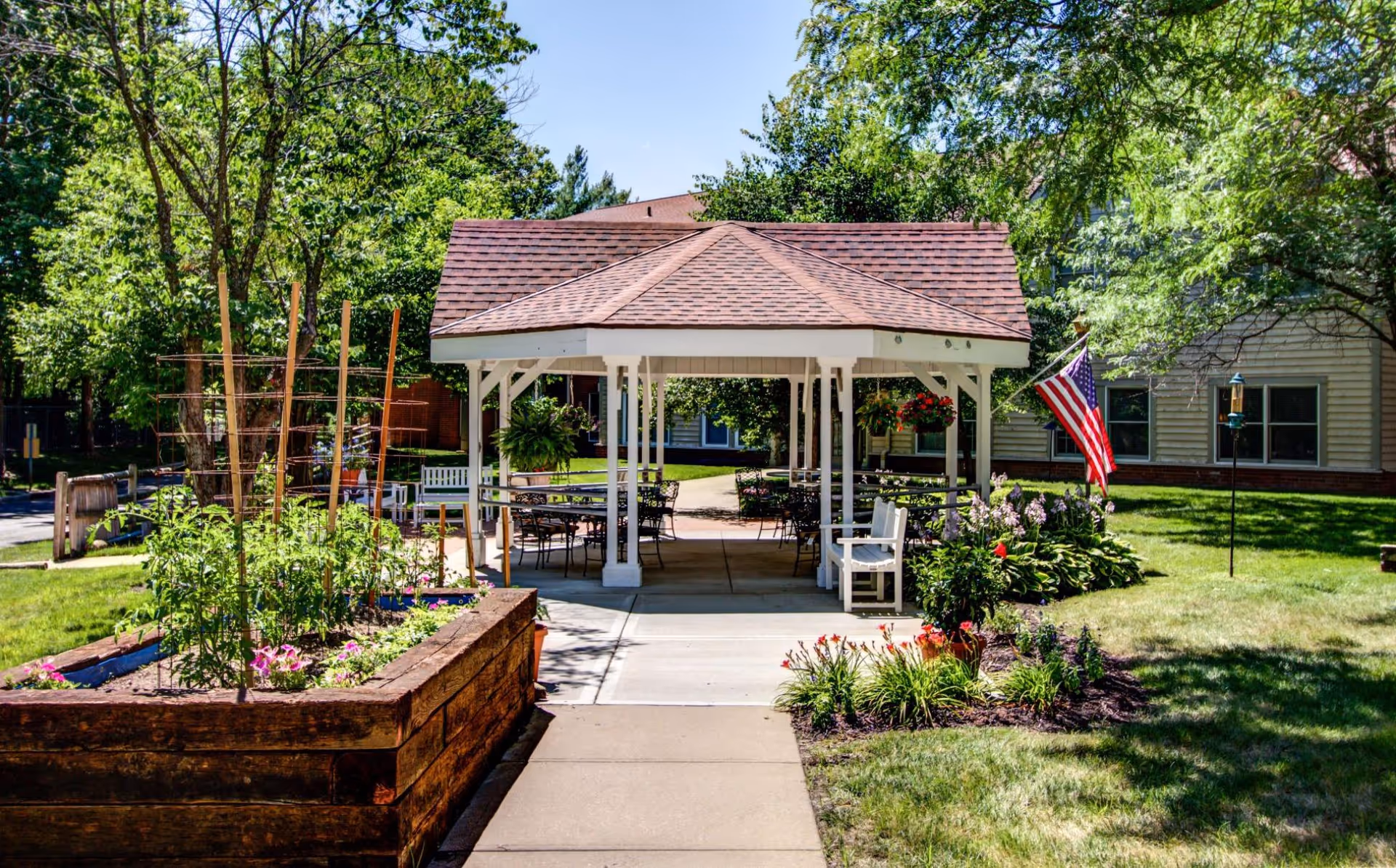 A garden area with a wooden raised planter box filled with plants and flowers on the left, a paved walkway leading to a white gazebo with a brown shingled roof. The gazebo has several chairs and tables underneath. There are trees and a building in the background, along with an American flag on the right side near some flower beds.