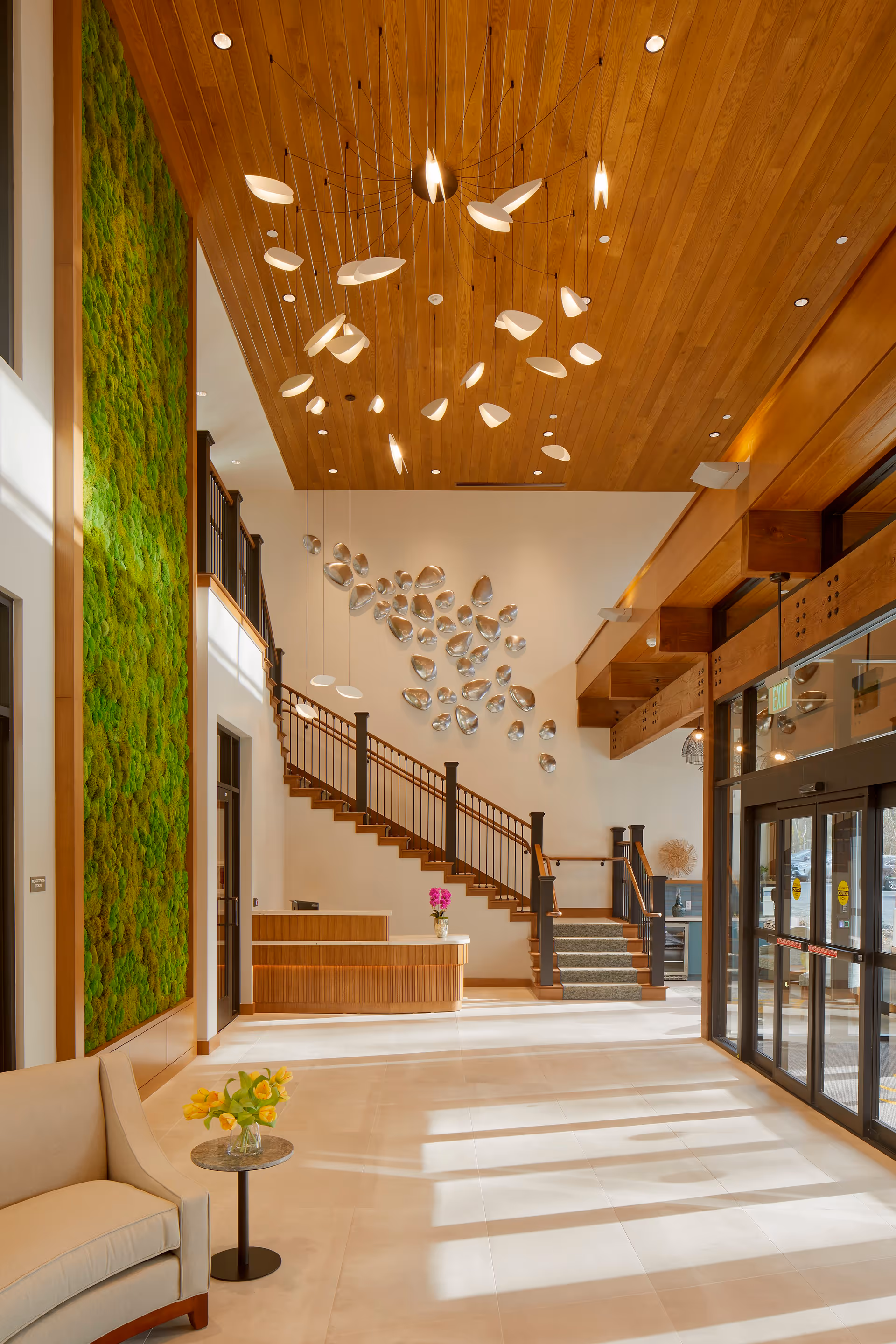 Bright and spacious lobby area with a wooden ceiling, modern hanging light fixtures, a green moss wall on the left, a wooden reception desk, and a staircase with dark railings leading to an upper floor. There is a beige sofa with a small round table holding yellow flowers, and large glass entrance doors on the right letting in natural light.
