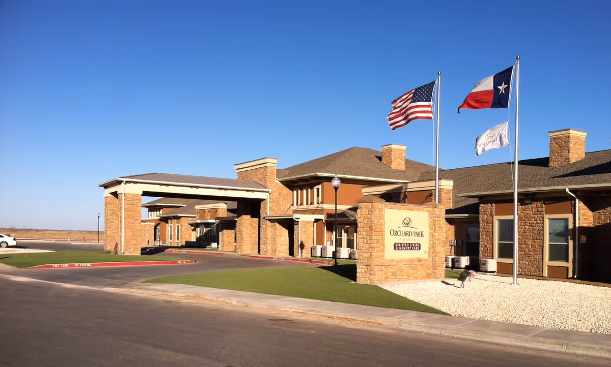 Front entrance of Orchard Park senior living building with flagpoles, a sign, and a drive-up canopy.